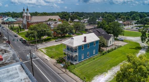 Aerial view of a blue house surrounded by green grass, with nearby buildings and trees under a partly cloudy sky.