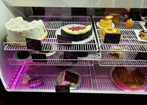 A display case filled with various cakes, including chocolate, red velvet, and lemon meringue, with labels.