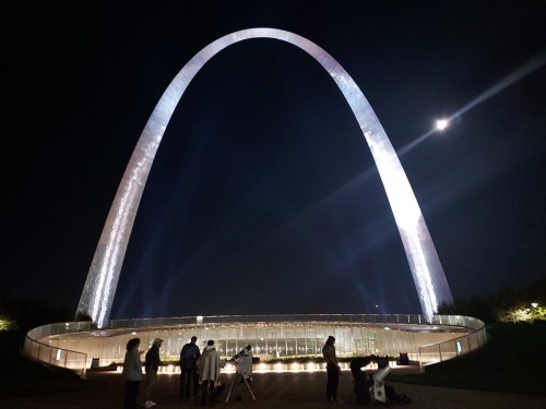A lit arch monument at night with people gathered below, observing the sky and a bright moon above.