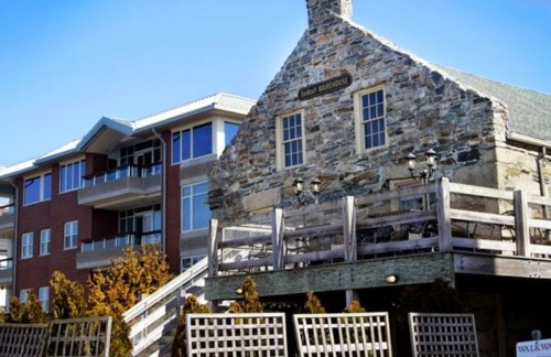 A stone building with a wooden deck in front, next to a modern multi-story apartment complex under a clear blue sky.