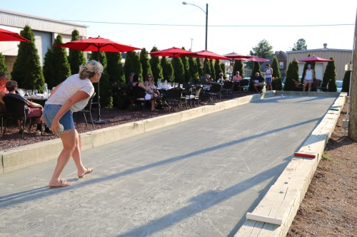 A woman prepares to throw a ball on a bocce court, with diners seated under red umbrellas in the background.