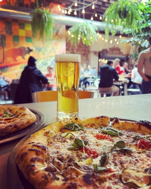 A close-up of two pizzas on a table with a glass of beer, set in a lively restaurant with colorful decor and patrons.
