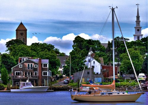 A scenic waterfront view featuring sailboats, trees, and buildings, with a clock tower and church steeple in the background.