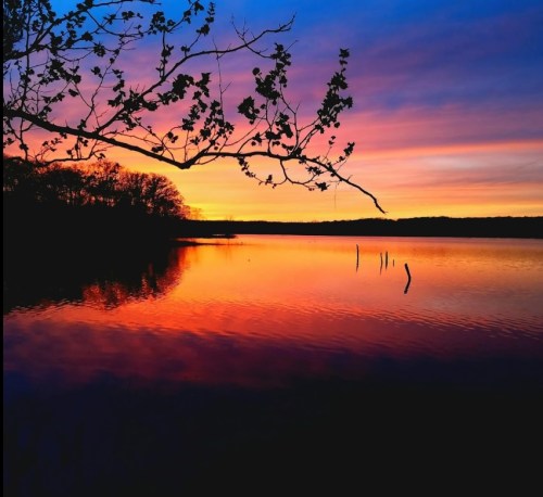A serene lake at sunset, reflecting vibrant orange and purple hues, with silhouetted trees along the shore.