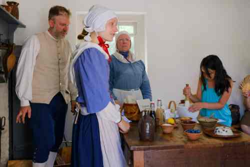 A historical scene with four people in period costumes, engaged in food preparation in a rustic kitchen.