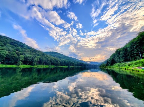 A serene lake reflecting clouds and mountains under a colorful sky, surrounded by lush greenery.