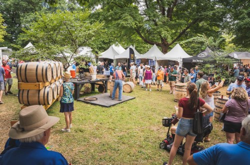 A lively outdoor festival scene with people enjoying food and drinks, surrounded by tents and wooden barrels.
