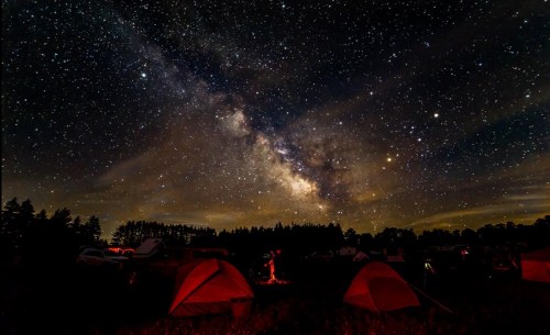 A starry night sky filled with the Milky Way over a campsite with tents and trees silhouetted against the horizon.