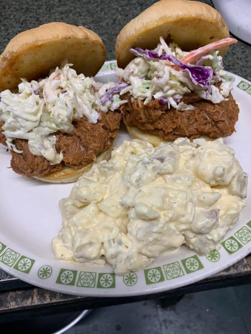 Two pulled pork sandwiches topped with coleslaw, served with a side of creamy potato salad on a plate.