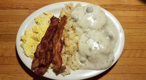A plate of breakfast featuring scrambled eggs, crispy bacon, hash browns, and biscuits smothered in creamy gravy.
