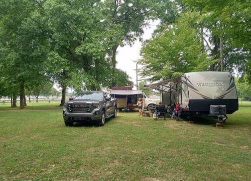 A campsite with a GMC truck and a Wilderness travel trailer surrounded by trees and grassy area.