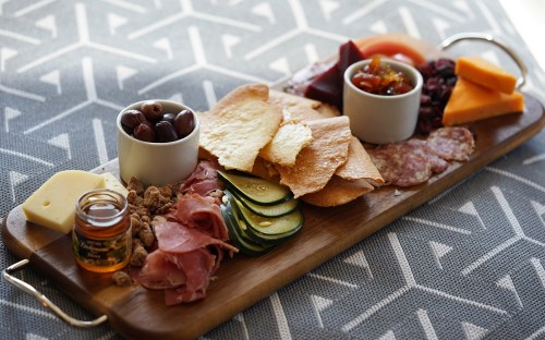 A wooden platter with assorted meats, cheeses, pickles, olives, chips, and two small bowls of condiments.