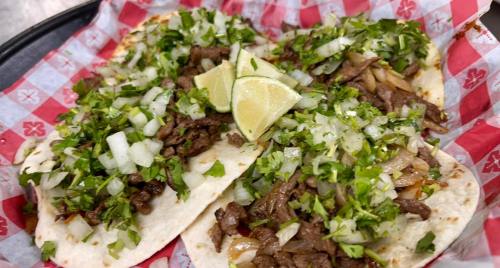 A close-up of three tacos filled with beef, topped with chopped onions, cilantro, and lime wedges on a red checkered paper.