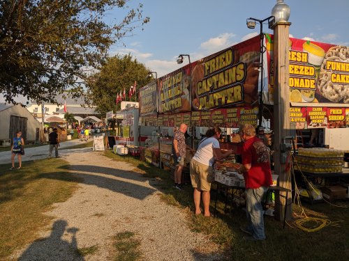 A lively outdoor food market with colorful stalls selling fried foods and treats, surrounded by trees and people enjoying the fair.