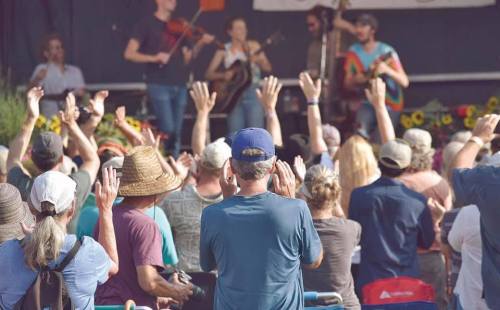 A crowd with raised hands enjoys a live music performance on stage, surrounded by greenery and bright flowers.