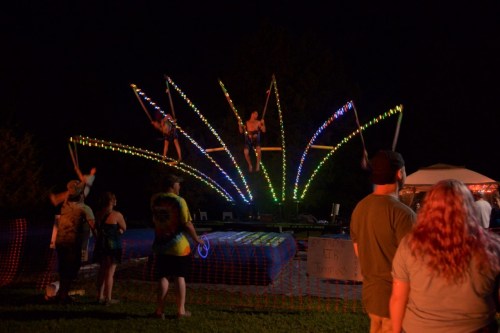 Three performers on a colorful, illuminated trampoline at night, with spectators watching nearby.