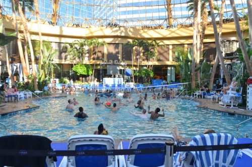 A lively indoor pool scene with people swimming and playing, surrounded by palm trees and lounge chairs.
