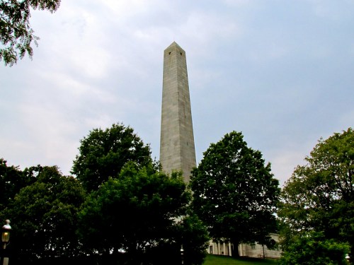 A tall stone obelisk surrounded by trees under a cloudy sky.