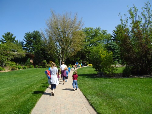 A group of children and adults walking along a paved path in a sunny park with green grass and trees.