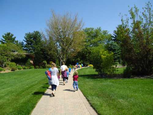 A group of children and adults walking along a paved path in a sunny park with green grass and trees.