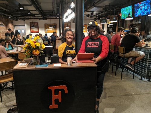 Two smiling staff members stand behind a counter at a busy restaurant, with flowers and a tablet in front of them.