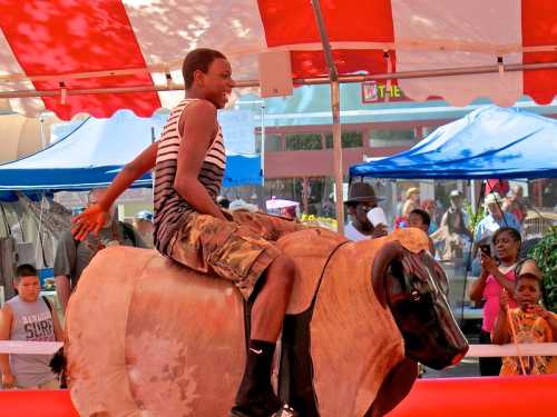 A young boy rides a mechanical bull at a fair, smiling as onlookers watch from the sidelines.