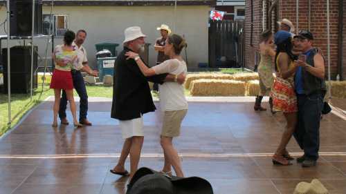 A lively outdoor dance scene with couples dancing on a wooden floor, surrounded by hay bales and festive decorations.