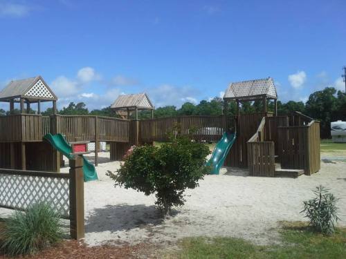 A wooden playground with slides and play structures, surrounded by sand and greenery under a blue sky.