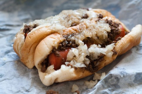A hot dog topped with seasoned ground beef and chopped onions, served in a soft bun on crinkled paper.