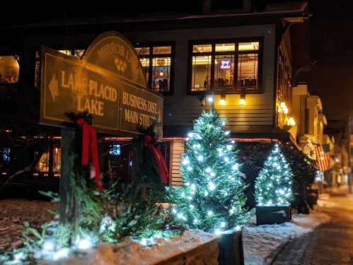 A cozy winter scene featuring a signpost and decorated Christmas trees with lights outside a building at night.
