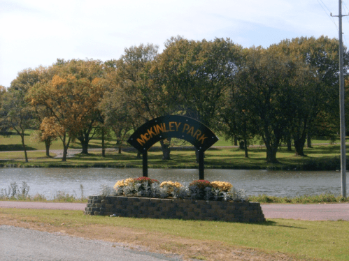 Sign for McKinley Park with colorful flowers in front, surrounded by trees and a lake in the background.