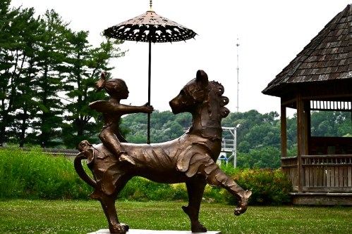 A bronze statue of a child holding an umbrella while riding a horse, set in a grassy area with trees in the background.