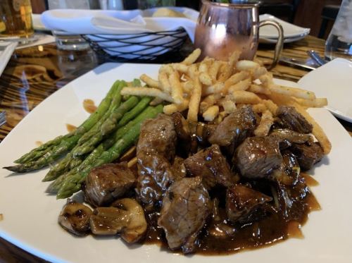 A plate of beef tips in sauce, served with asparagus and crispy fries. A copper mug is in the background.