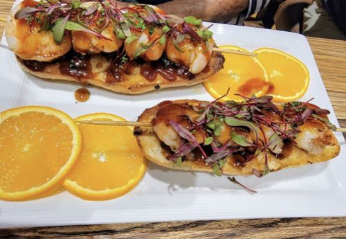 Two skewers of shrimp on toasted bread, drizzled with sauce, garnished with microgreens, and served with orange slices.