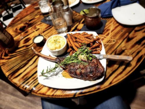 A large grilled steak with herbs, served with sweet potato fries and mashed potatoes on a wooden table.