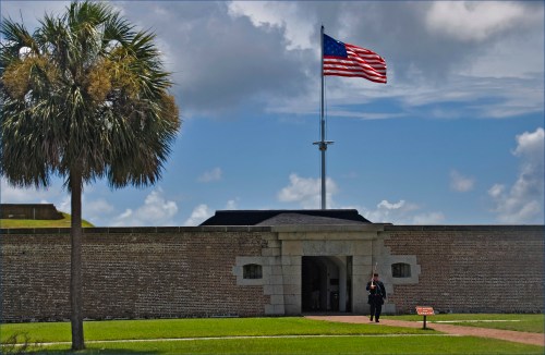 A soldier stands guard at a historic fort with an American flag flying above, surrounded by green grass and palm trees.