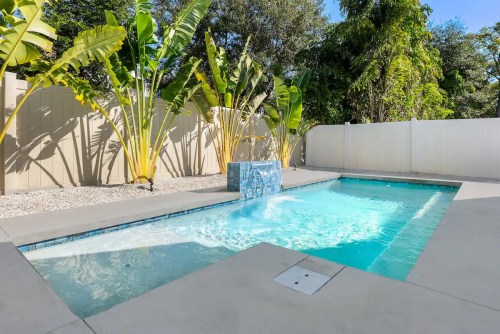 A serene backyard pool surrounded by tropical plants and a white fence, featuring a small water feature.