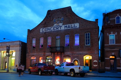 Historic concert hall at dusk, featuring a brick facade and colorful lights, with parked trucks in front.