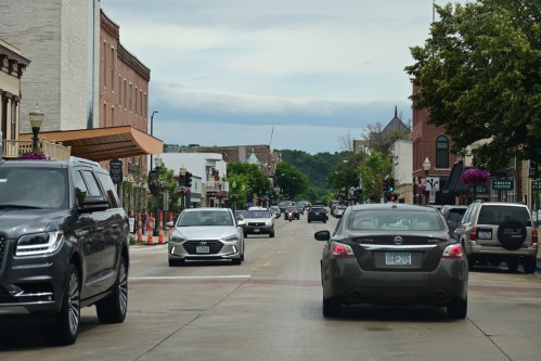 A busy street scene with cars driving, lined with shops and trees under a cloudy sky.
