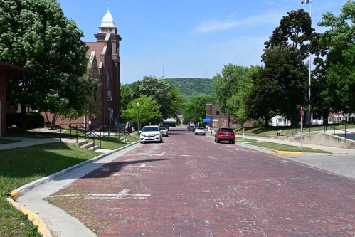 A quiet street lined with trees and brick buildings, featuring a brick road and parked cars under a clear blue sky.