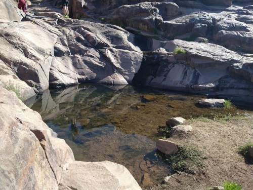 A rocky landscape featuring a shallow pool of water surrounded by large stones and patches of grass.