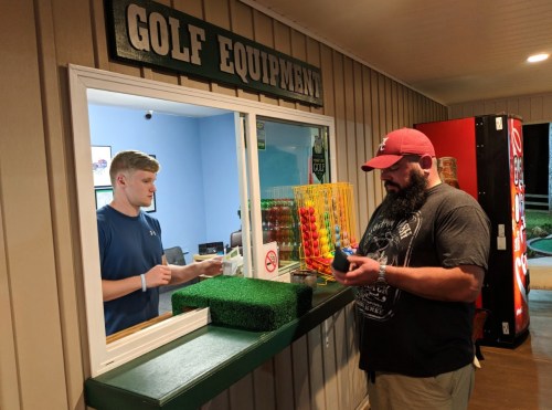 A man at a golf equipment counter interacts with a staff member while holding a colorful item.