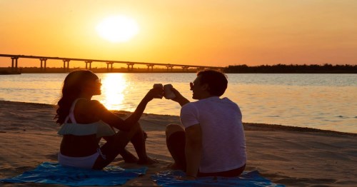 A couple toasting drinks at sunset on the beach, with a bridge in the background and a serene water view.