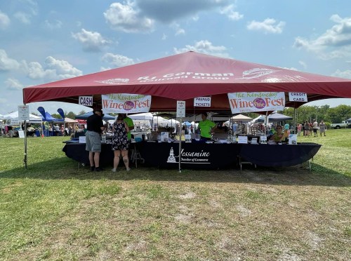 A tent at a festival with a sign for "Kentucky Wine Fest," featuring vendors and attendees in a grassy field.