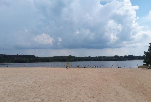 A sandy beach with a calm lake, under a cloudy sky, and people enjoying the water in the distance.