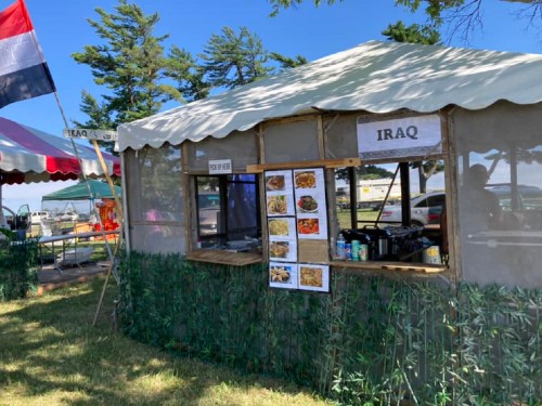 A food stall labeled "Iraq" with a menu display, set up outdoors under a tent with trees in the background.