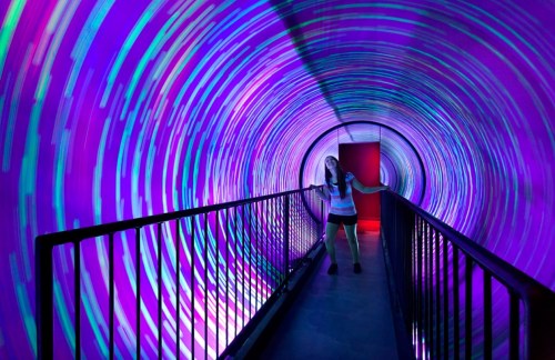 A person stands in a colorful, illuminated tunnel with swirling patterns, surrounded by vibrant purple and blue lights.