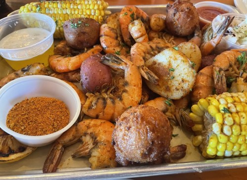 A seafood platter featuring shrimp, corn, potatoes, and dipping sauces on a tray, garnished with herbs.