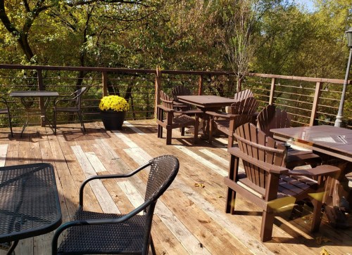 A wooden deck with several outdoor tables and chairs, surrounded by trees and autumn foliage.