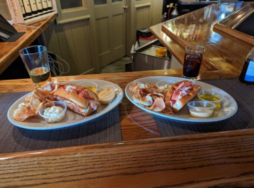 Two plates of lobster rolls with sides of coleslaw and dipping sauces, accompanied by drinks on a wooden bar.